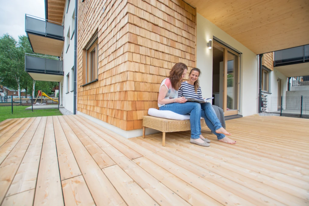 Zwei Frauen sitzen auf einer Holzterrasse vor einem Wohngebäude in Massivholzbauweise in Niedernsill.