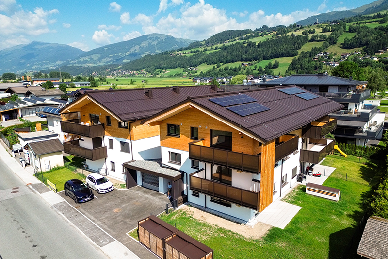 Luftaufnahme eines modernen Wohngebäudes in Mittersill mit Holzfassade, Balkonen, Photovoltaikanlage und umliegender Berglandschaft.